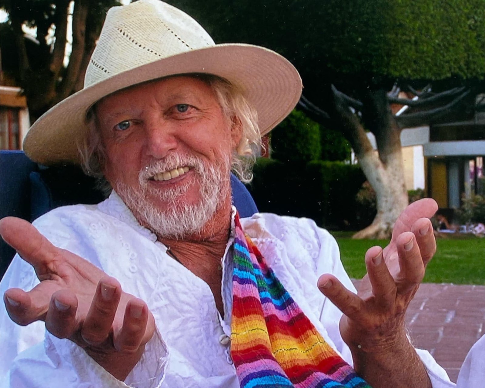 Edwin Riley, seated outdoors in a straw hat, hands open mid-story.